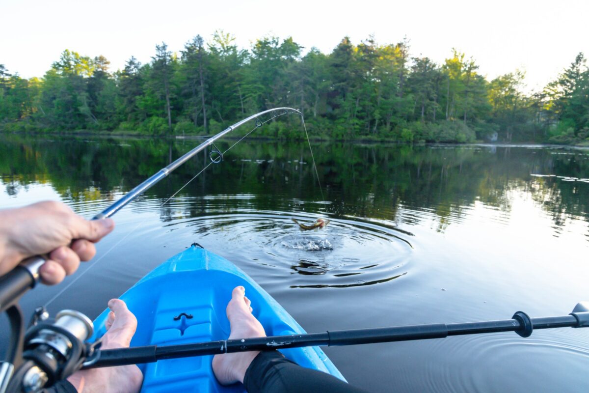 Angler holding a fishing rod from a blue kayak as a hooked fish breaks the surface of a lake