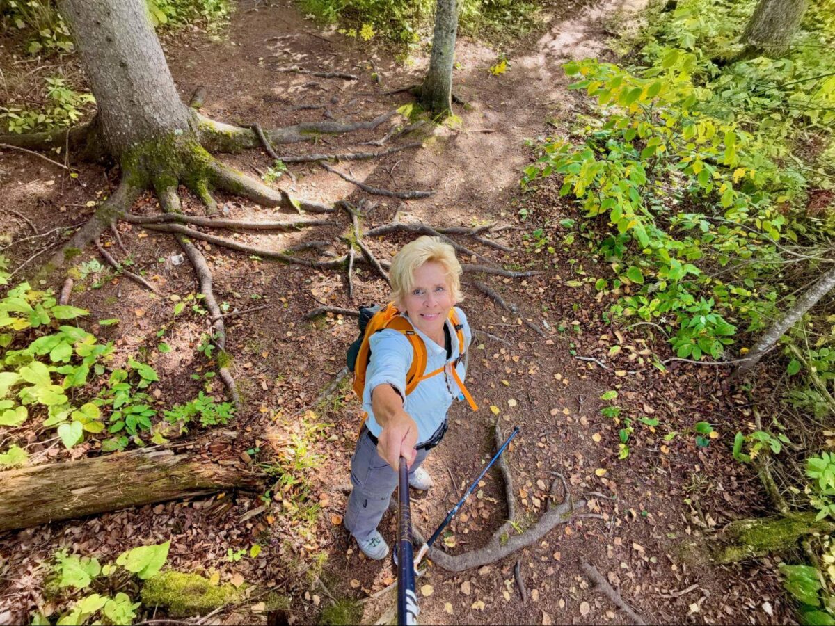 Krissie Mason holding a selfie stick on a forest trail with exposed tree roots and uneven ground