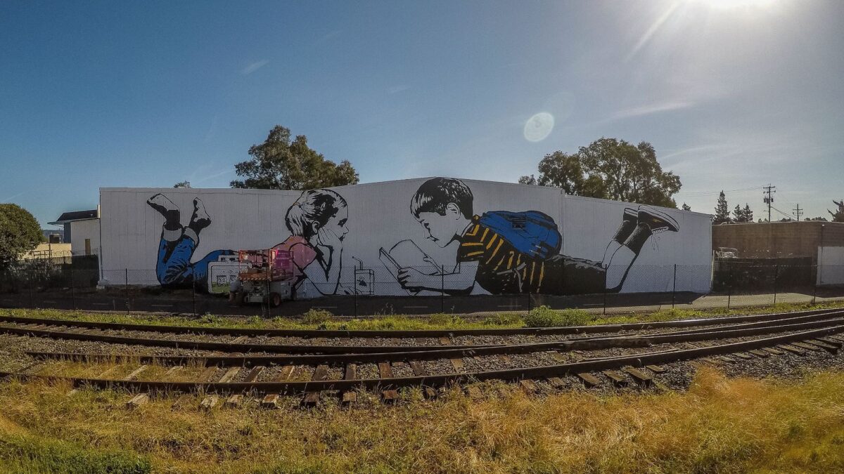 A large mural depicts two children lying on their stomachs, one reading a book, on a building beside railroad tracks in Napa.